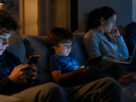 Family members sitting together in a living room at night while each person uses a different digital device, illustrating concerns about excessive screen time and digital disconnection in modern households.