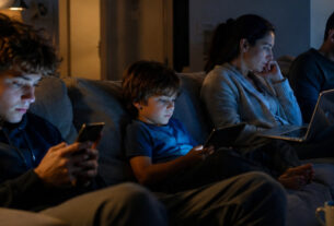 Family members sitting together in a living room at night while each person uses a different digital device, illustrating concerns about excessive screen time and digital disconnection in modern households.